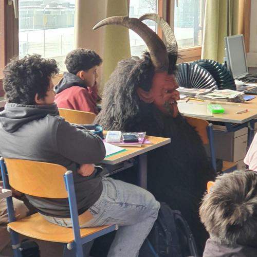 Das Bild zeigt einen Krampus zwischen den Schülern sitzend