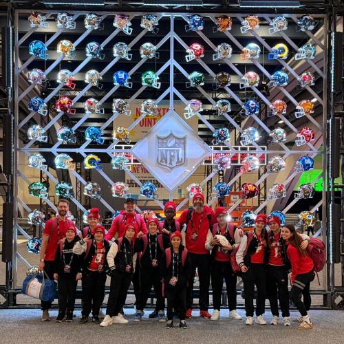 Das Bild zeigt das Team vor einer NFL Wand mit Football-Helmen